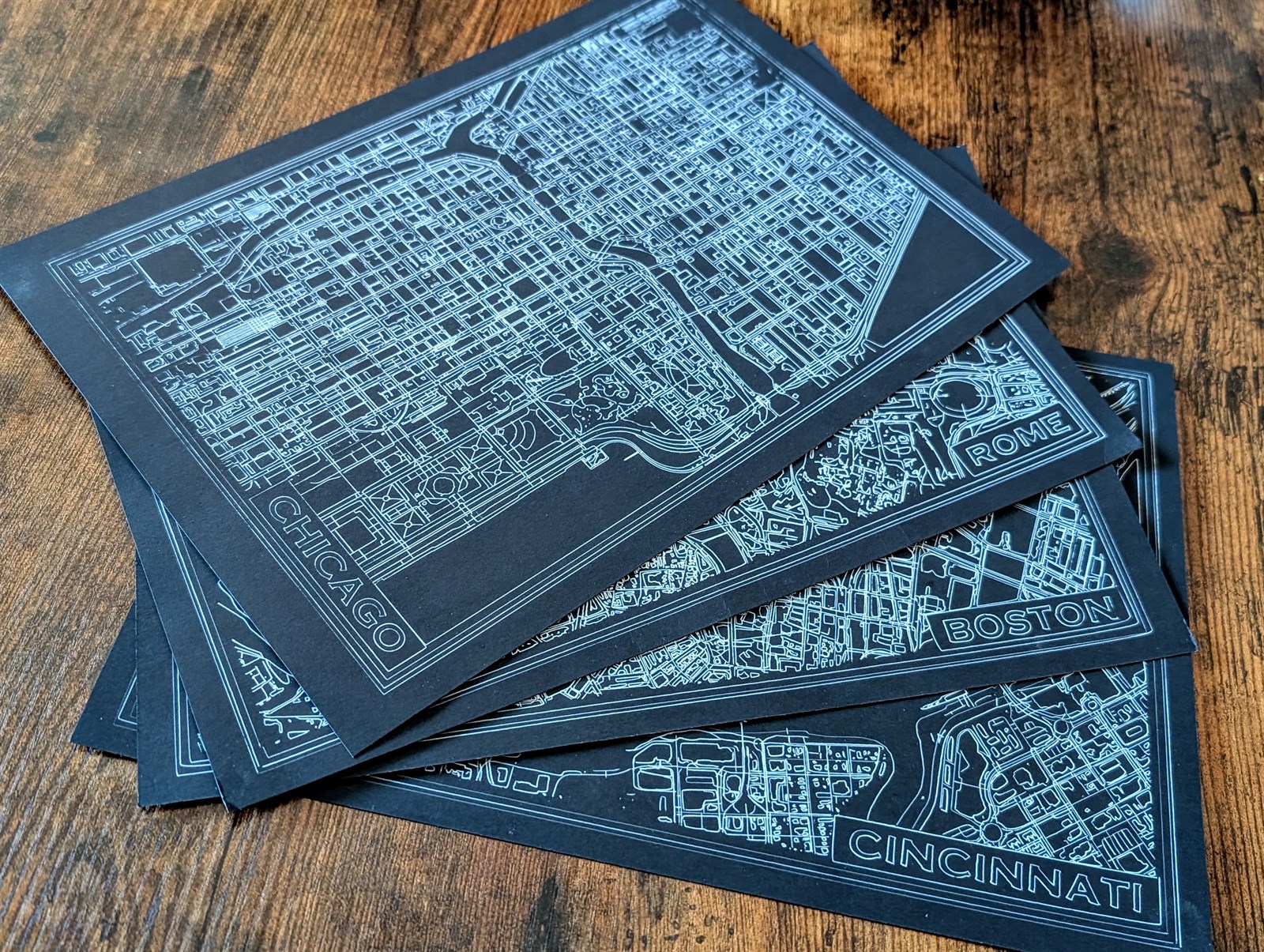 Finished laser-engraved city maps stacked on a wooden table, including Chicago, Rome, Boston, and Cincinnati